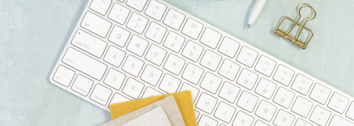 White keyboard with different colour fabric samples on it. Keyboard is surrounded with a turquoise coloured glass with lemon on the side, a candle, scissors, note pad and pen and headphones.