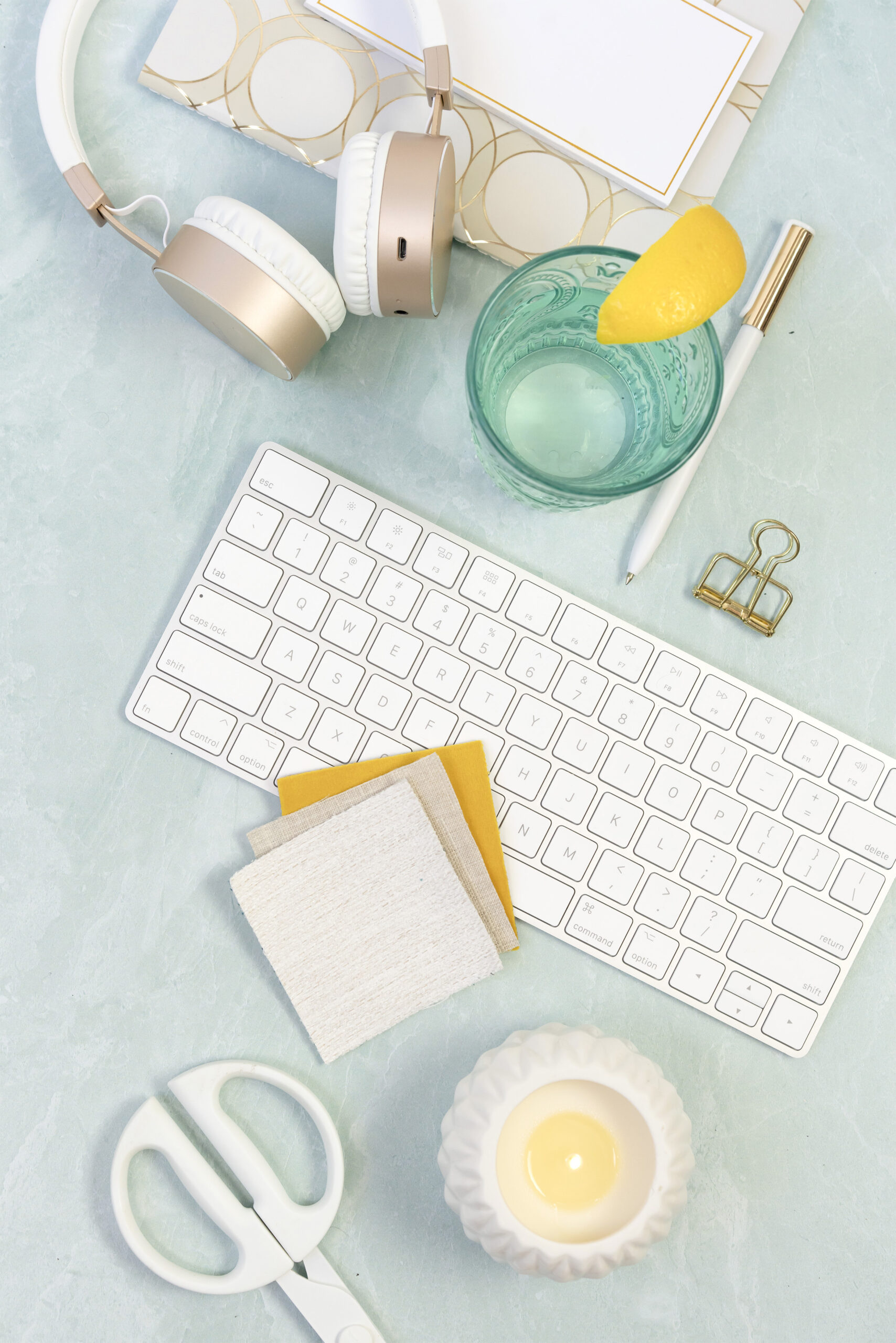 White keyboard with different colour fabric samples on it. Keyboard is surrounded with a turquoise coloured glass with lemon on the side, a candle, scissors, note pad and pen and headphones.